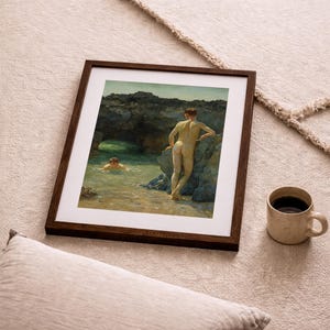 Op de afbeelding: Een ingelijste prent van een klassiek schilderij met twee naakte figuren bij een rotsachtige kustlijn. E&eacute;n figuur staat op de oever, de ander zwemt in het water. Een beige mok met donkere vloeistof staat in de buurt.