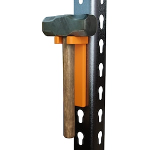 May include: A black and orange hammer holder attached to a black metal pegboard. The holder is designed to hold a hammer with a wooden handle.