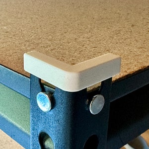 May include: A white plastic corner protector is attached to a black metal shelf unit. The corner protector is designed to protect the shelf from damage.