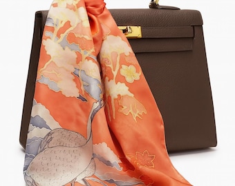 May include: A brown leather handbag with a top handle and gold-tone hardware. An orange silk scarf with a crane and floral design is draped over the bag. The scarf features a detailed illustration with shades of orange, white, and blue.