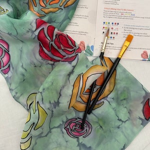 May include: A silk scarf with a green background and a floral design featuring roses in shades of red, orange, yellow, and pink. The scarf is folded and three paintbrushes are lying on top of it. The text on the paper reads "For Silk Scarf Owners".