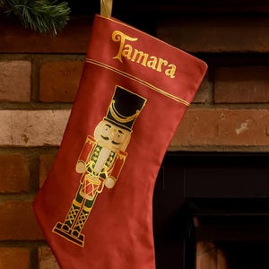 以下が含まれることがあります： 金色のループが付いた赤いクリスマスストッキング。名前「Tamara」が金色の文字で刺繍されています。赤、黒、金、緑色のくるみ割り人形の兵士のデザインが刺繍されています。クリスマスの装飾。