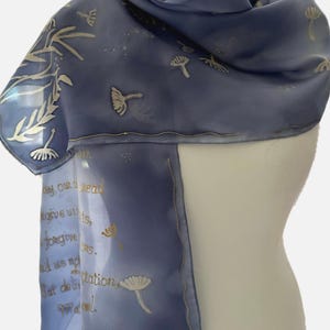 May include: A light blue silk scarf with a white dandelion design and gold lettering. The text reads "Give us this day our daily bread, forgive us our trespasses, as we forgive those who trespass against us. And lead us not into temptation, but deliver us from evil."