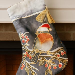 以下が含まれることがあります： サンタ帽をかぶった鳥が赤い実のついた枝にとまっているクリスマスストッキング。ストッキングはグレーの背景、白いフェイクファーのカフス、金のタッセルが付いています。