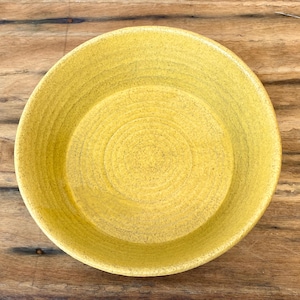 May include: A round, yellow ceramic bowl with a textured surface. The bowl has a wide rim and a shallow center, with concentric circles forming the base. The bowl is likely handmade and suitable for serving food.