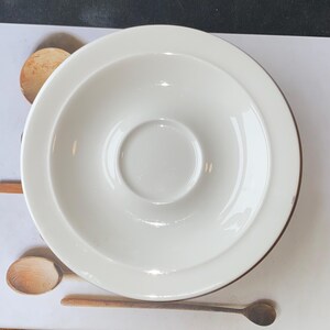 Op de afbeelding: Een witte keramische schotel met een verzonken midden, ontworpen om een kopje in te plaatsen. De schotel heeft een brede rand en een licht verhoogde rand. Houten lepels zijn ook zichtbaar.