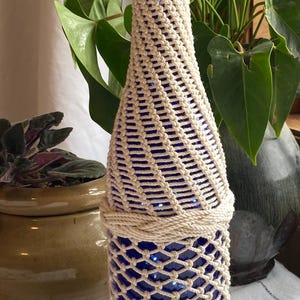 May include: A cobalt blue glass bottle encased in a decorative off-white macrame cord netting. The intricate knotted rope design covers the neck and body of the bottle, featuring horizontal braided rope bands at the top, middle, and base sections.