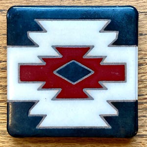 May include: Square ceramic coaster with a geometric design. The coaster features a central red and navy blue diamond shape, surrounded by white and navy blue zig-zag patterns. The coaster has a glossy finish.