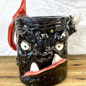 May include: Black ceramic mug with a monster face design. The mug features white eyes, large teeth, and a red handle and lip. The mug has a glossy finish and a textured surface.