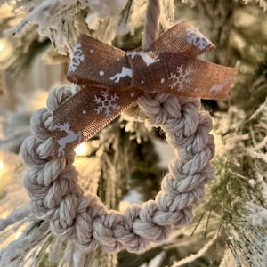 May include: A beige rope wreath ornament with a brown ribbon with white snowflakes and reindeer printed on it. The ornament is hanging on a snowy artificial Christmas tree.