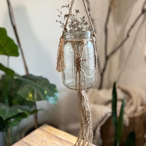 May include: A clear glass jar with dried flowers inside is suspended from a macrame plant hanger made of natural jute rope. The hanger has a long tassel at the bottom.
