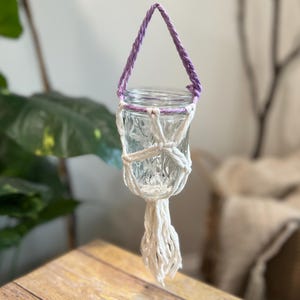 Puede incluir: Un macetero de macramé blanco con un lazo de suspensión morado, que sostiene un tarro de vidrio transparente.