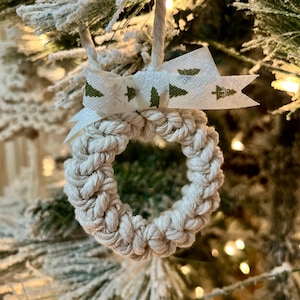 May include: A small, white, crocheted wreath ornament with a white ribbon with green tree print. The ornament is hanging on a snowy Christmas tree.