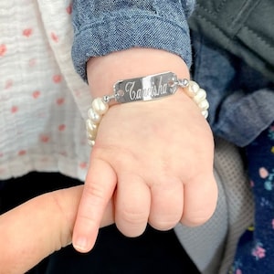 May include: A silver bracelet with a personalised nameplate that reads "Carieisha" and white pearl beads.