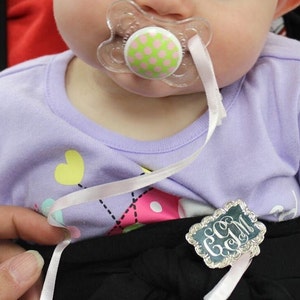 May include: A baby wearing a purple shirt with a pink and green pacifier and a silver monogrammed tag attached to a white ribbon.