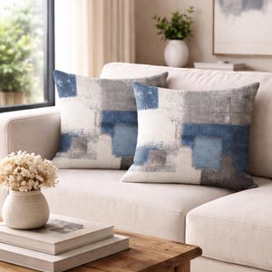 May include: Two square throw pillows with a modern abstract design. The pillows feature a pattern of blue, gray, and white blocks, creating a textured, artistic look. They are placed on a cream-colored sofa, adding a touch of contemporary style to the living space.