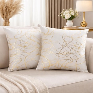 Puede incluir: Dos cojines blancos con diseños de líneas doradas abstractas sobre un sofá beige. Los cojines tienen un aspecto suave y texturizado. Las líneas doradas crean un aspecto moderno y elegante, adecuado para la decoración del hogar.
