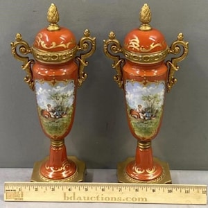 K&ouml;nnte beinhalten: Ein Paar antike Porzellanurnen mit goldenen Akzenten und bemalten Szenen. Die Urnen sind orange mit goldenen Verzierungen und zeigen eine pastorale Szene mit Figuren in einer Landschaft.