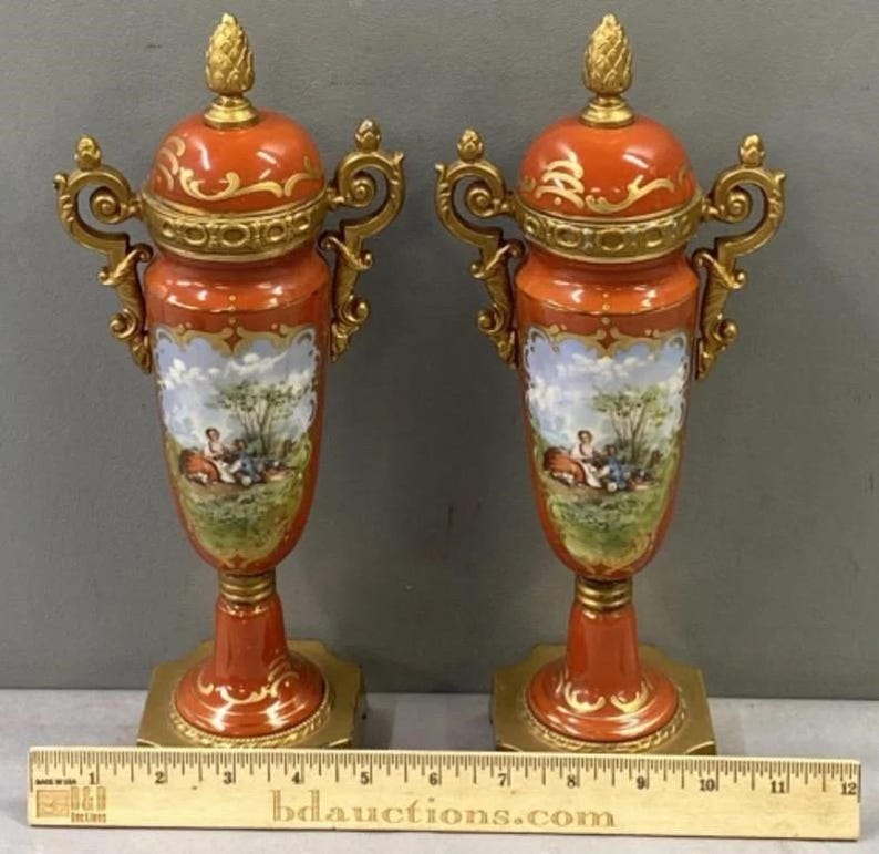 K&ouml;nnte beinhalten: Ein Paar antike Porzellanurnen mit goldenen Akzenten und bemalten Szenen. Die Urnen sind orange mit goldenen Verzierungen und zeigen eine pastorale Szene mit Figuren in einer Landschaft.