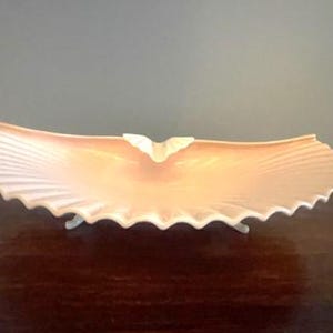 May include: A pale pink ceramic bowl with a scalloped edge and three legs. The bowl is shaped like a seashell and has a delicate, feminine design.