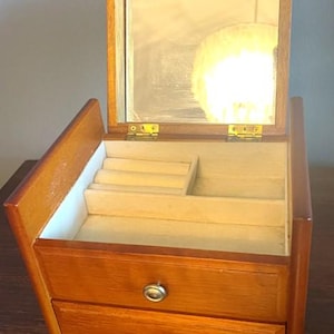 May include: A wooden jewelry box with a hinged lid and a mirror inside. The box has two drawers with brass knobs. The interior is lined with beige felt.