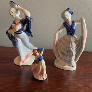 May include: Three porcelain figurines of women in blue and white dresses with pink accents. The figurines are in various poses, with one holding a hat.
