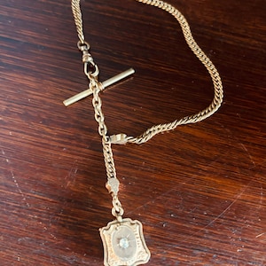 Pode incluir: Uma corrente de relógio de bolso de ouro com um pendente decorativo. A corrente é feita de um design de corda torcida e tem um fecho em T. O pendente é um medalhão em forma de escudo com um único diamante no centro.