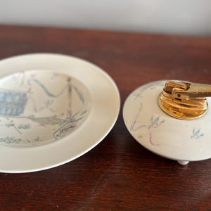 Pode incluir: Uma tigela de cerâmica branca e um isqueiro com a parte superior dourada. A tigela tem uma borda larga e um desenho pintado em azul, cinza e verde. O isqueiro tem formato oval com uma alavanca dourada.