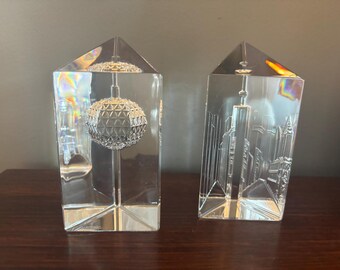 May include: Two clear glass paperweights with 3D etched designs. One paperweight features a depiction of the New York City skyline, while the other features a depiction of the New York City ball drop.