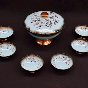 May include: A vintage serving set with a lidded bowl and six small bowls. The set features a light blue speckled glaze with brown and gold accents. The bowls are set on copper-colored bases.