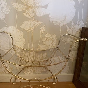 May include: A gold metal stool with a curved back and a wire mesh seat. The stool is in front of a wall with a floral pattern.