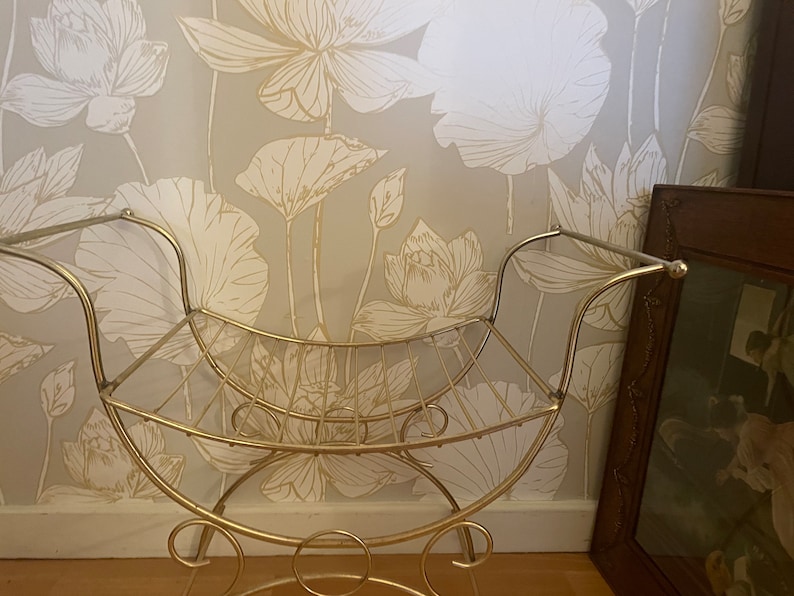 May include: A gold metal stool with a curved back and a wire mesh seat. The stool is in front of a wall with a floral pattern.