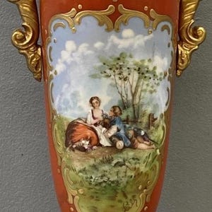 K&ouml;nnte beinhalten: Eine rote und goldene Porzellanvase mit einer gemalten Szene von zwei Figuren in einer Landschaft. Die Vase hat verzierte goldene Akzente und ein zartes Blumenmuster.