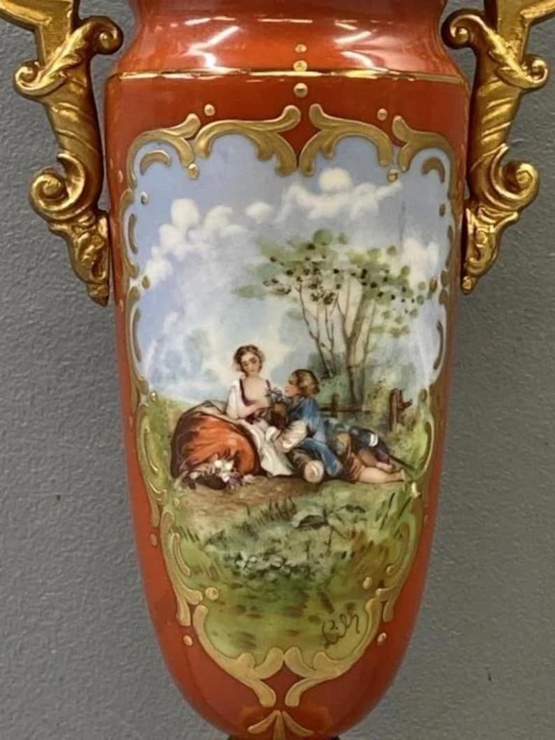 K&ouml;nnte beinhalten: Eine rote und goldene Porzellanvase mit einer gemalten Szene von zwei Figuren in einer Landschaft. Die Vase hat verzierte goldene Akzente und ein zartes Blumenmuster.