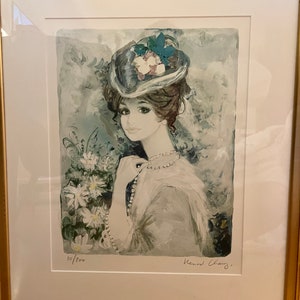 May include: A framed print of a woman wearing a blue hat with flowers and a pearl necklace. The print is signed "Bernard Charoy" and numbered "10/200".