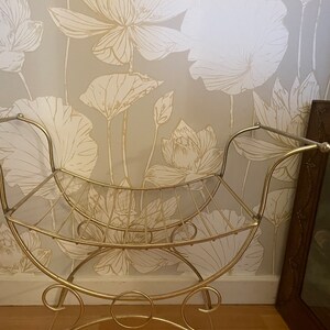 May include: A gold metal stool with a curved seat and a shelf underneath. The stool is in front of a wall with a floral pattern.