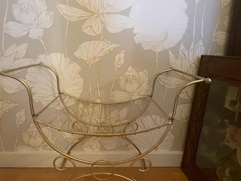 May include: A gold metal stool with a curved seat and a shelf underneath. The stool is in front of a wall with a floral pattern.