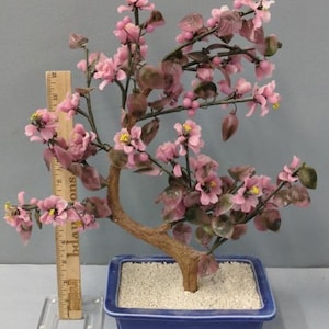 Puede incluir: Un bonsái de cristal rosa con delicadas flores y hojas. El árbol está plantado en una maceta rectangular azul llena de guijarros blancos. El tronco es marrón y las flores tienen centros amarillos.