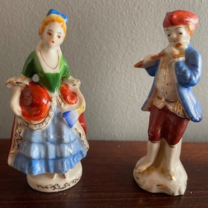 May include: A pair of vintage porcelain figurines depicting a woman in a blue and green dress holding a red ball and a man in a blue jacket playing a flute.