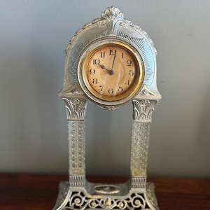May include: A silver-toned, ornate, decorative clock with a round, cream-colored face and black hands. The clock has a decorative, arched top and a base with intricate details.