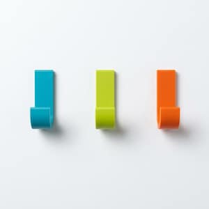 Könnte beinhalten: Drei J-förmige Wandhaken, die in einer horizontalen Reihe angeordnet sind. Der linke Haken ist leuchtend blau, der mittlere Haken ist limettengrün und der rechte Haken ist orange. Jeder Haken hat eine flache Rückplatte und eine gebogene Stütze.