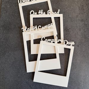 Könnte beinhalten: Vier weiße, rechteckige Fotorahmen in Form von Sofortbildern. Jeder Rahmen trägt oben einen individuellen Schriftzug in Schreibschrift: Wild & Free, On the Road, Happy Camper und Campfire Days. Die Rahmen sind in einer Schichtung angeordnet.
