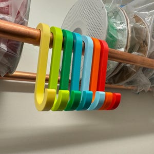 Peut inclure: Une série de crochets en forme de S dans une palette de couleurs arc-en-ciel vives, incluant jaune, vert lime, vert forêt, bleu sarcelle, bleu clair, orange et rouge, suspendus à une tige métallique horizontale cuivrée, finition mate.