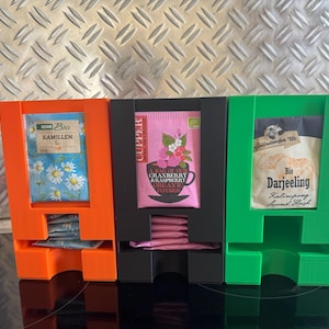 Könnte beinhalten: Drei vertikale Teebeutelspender in Orange, Schwarz und Grün. Der orangefarbene Halter enthält Kamillen-Tee, der schwarze Halter enthält Cranberry & Raspberry Bio-Aufguss und der grüne Halter enthält Bio-Darjeeling-Teebeutel.