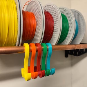 Könnte beinhalten: Fünf S-förmige Kunststoffhaken in Gelb, Orange, Rot, Grün und Blau hängen an einem horizontalen Kupferrohr. Hinter den Haken befinden sich fünf Spulen mit 3D-Drucker-Filament in passenden Farben auf einem Regal mit schwarzen Halterungen.