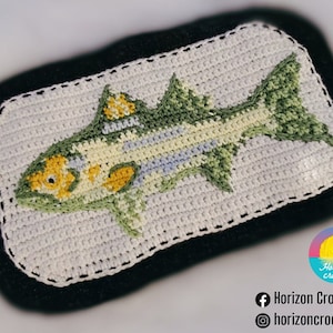Peut inclure: Un motif de poisson crocheté dans les tons de vert, jaune et bleu, sur fond blanc avec une bordure noire. Le poisson est détaillé avec des écailles et des nageoires, mettant en valeur un artisanat fait à la main. Il s'agit probablement d'un objet décoratif ou d'une œuvre d'art.