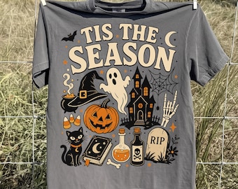 Może przedstawiać: Ciemnoszary t-shirt z napisem TIS THE SEASON kremowymi literami. Grafika przedstawia czarny kapelusz czarownicy, białego ducha, dynię, czarnego kota, księgę zaklęć, buteleczki z miksturami, nawiedzony dom oraz nagrobek z napisem RIP.