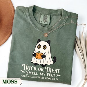 A funny Halloween graphic t-shirt featuring a cute black cat wearing a classic ghost sheet costume and holding a small pumpkin. The vintage style text reads TRICK OR TREAT SMELL MY FEET GIVE ME SOMETHING GOOD TO EAT with small flying bats. Perfect retro spooky season apparel and a funny cat lover gift.