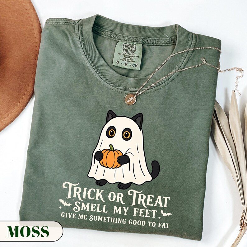 A funny Halloween graphic t-shirt featuring a cute black cat wearing a classic ghost sheet costume and holding a small pumpkin. The vintage style text reads TRICK OR TREAT SMELL MY FEET GIVE ME SOMETHING GOOD TO EAT with small flying bats. Perfect retro spooky season apparel and a funny cat lover gift.