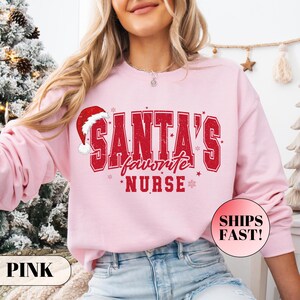 Puede incluir: Una sudadera de manga larga de color rosa claro con texto rojo desgastado de estilo universitario que dice Santa's favorite NURSE. El diseño incluye un gráfico de gorro de Papá Noel rojo y blanco sobre la letra S y estrellas.